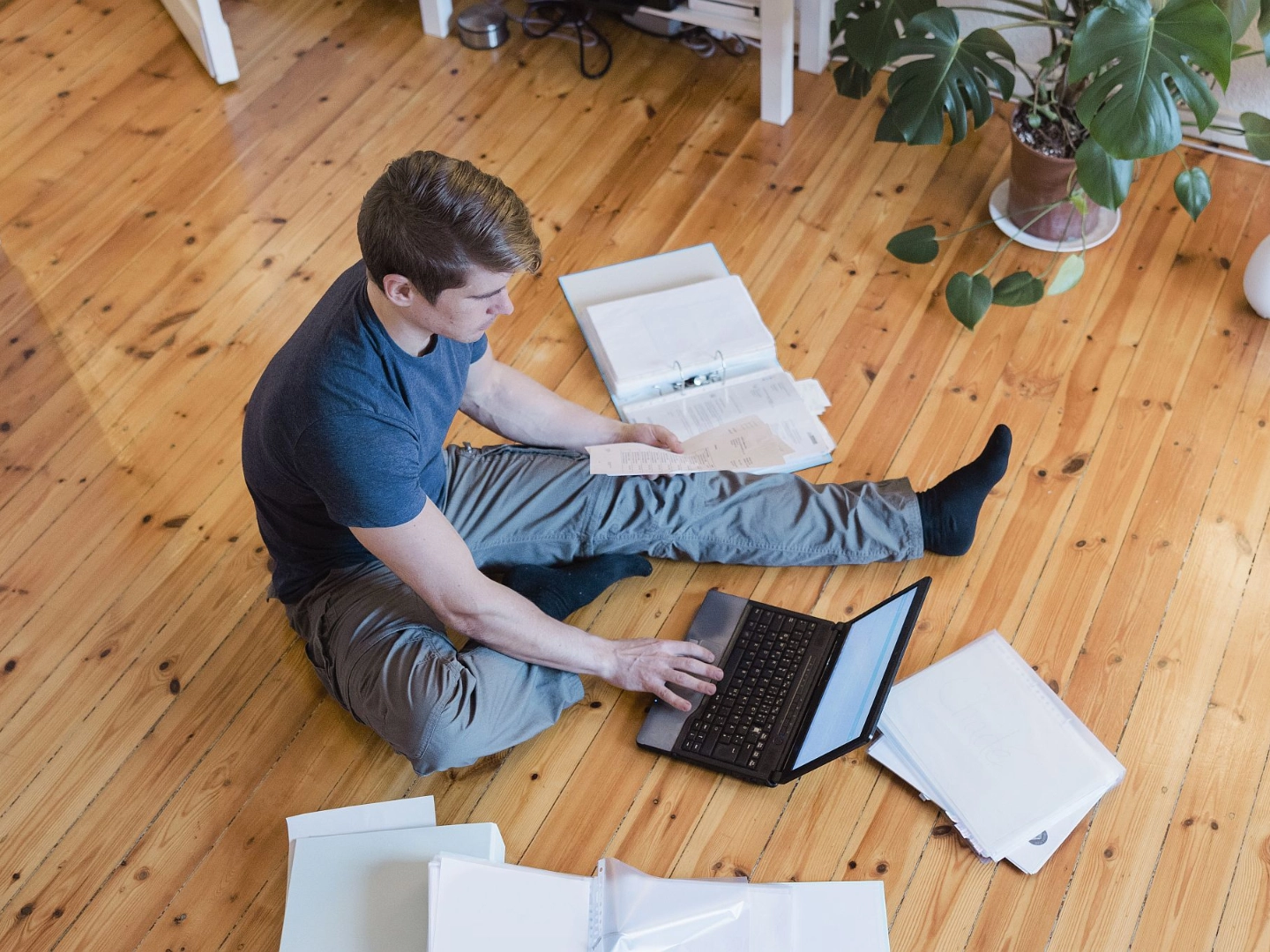 Junger Mann sitzt mit Notebook auf Fußboden und sortiert Papier und Akten
