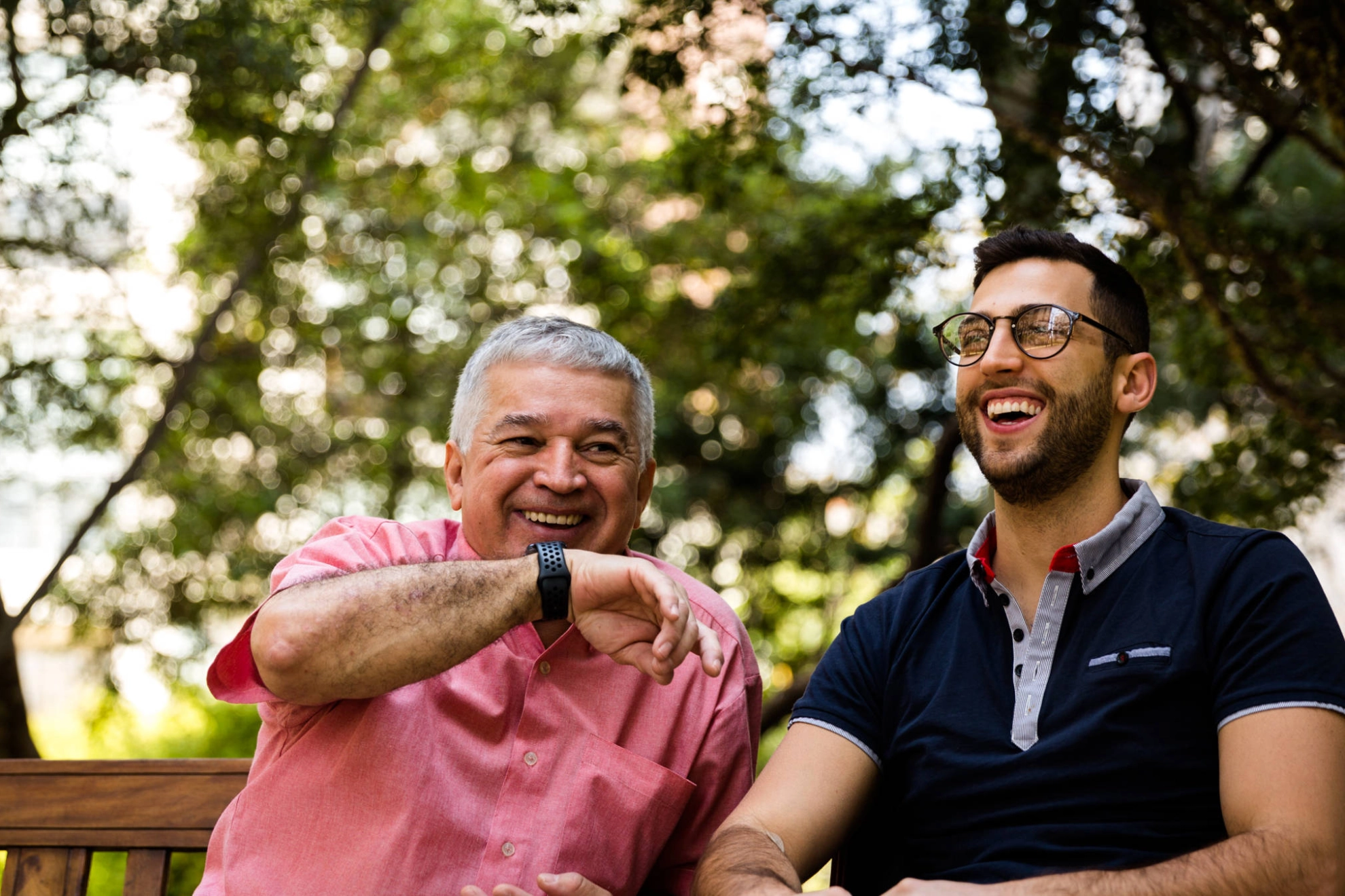 Ein junger und ein älterer Mann sitzen lachend nebeneinander auf einer Bank.