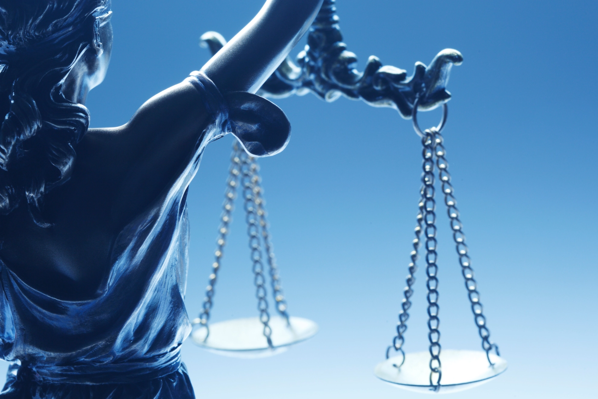 Ein Bild von Justizia als Bronzestatue vor blassblauem Hintergrund, auf dem sie eine Waage hält