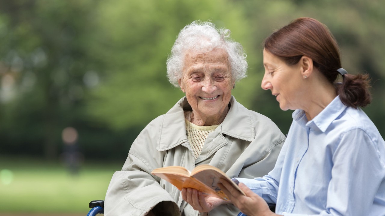 Eine Seniorin im Rollstuhl und ihre Tochter schauen in einem Park gemeinsam in ein Buch.
